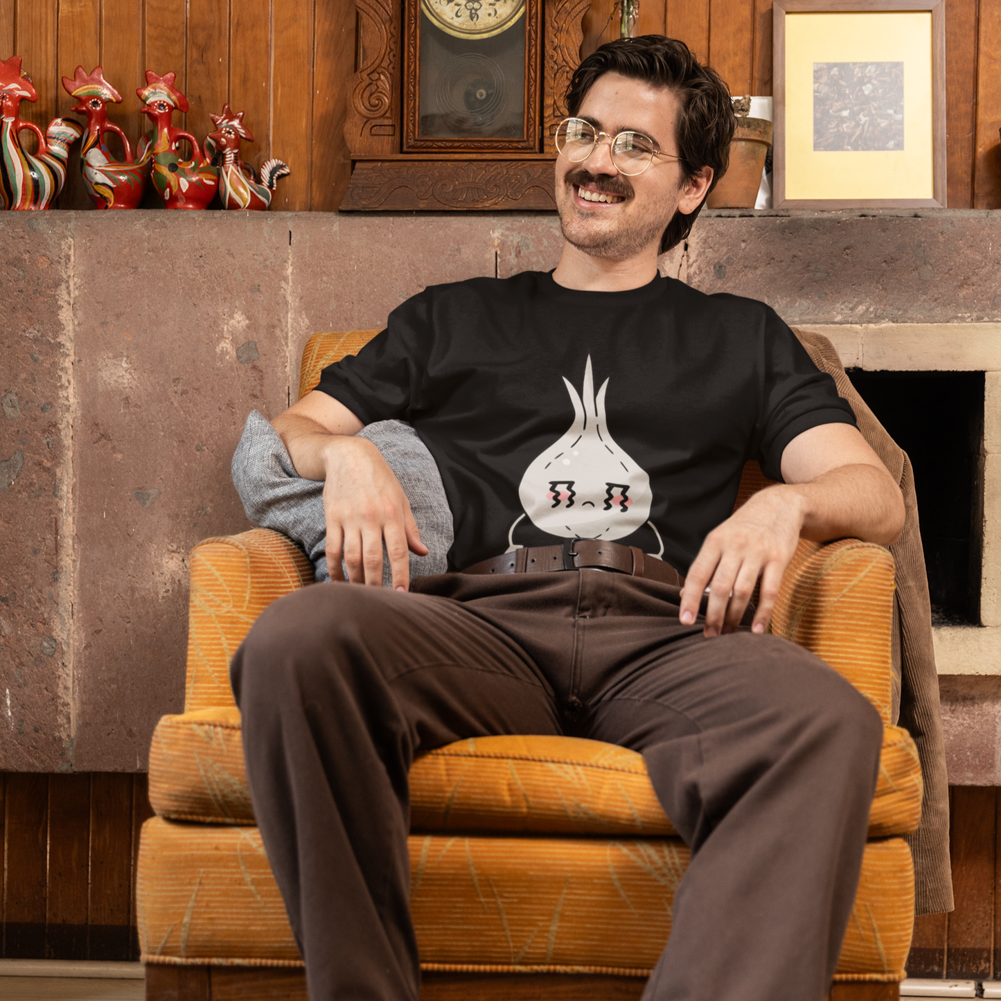 Man sitting in an armchair wearing a black t-shirt with a graphic design, in a room with wooden walls and decorative items.