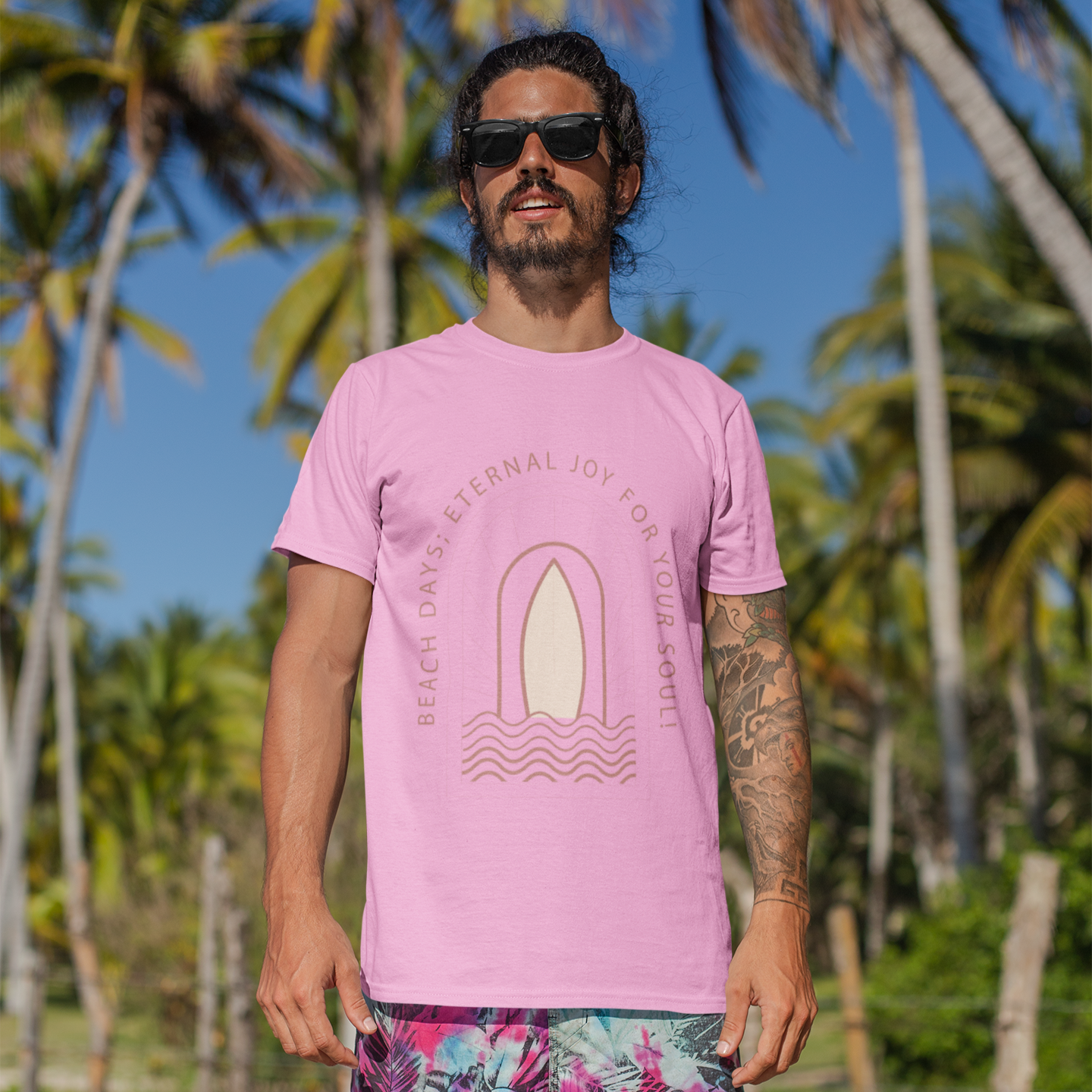 Man wearing a pink t-shirt with a graphic design and colorful shorts standing on a beach with palm trees in the background.