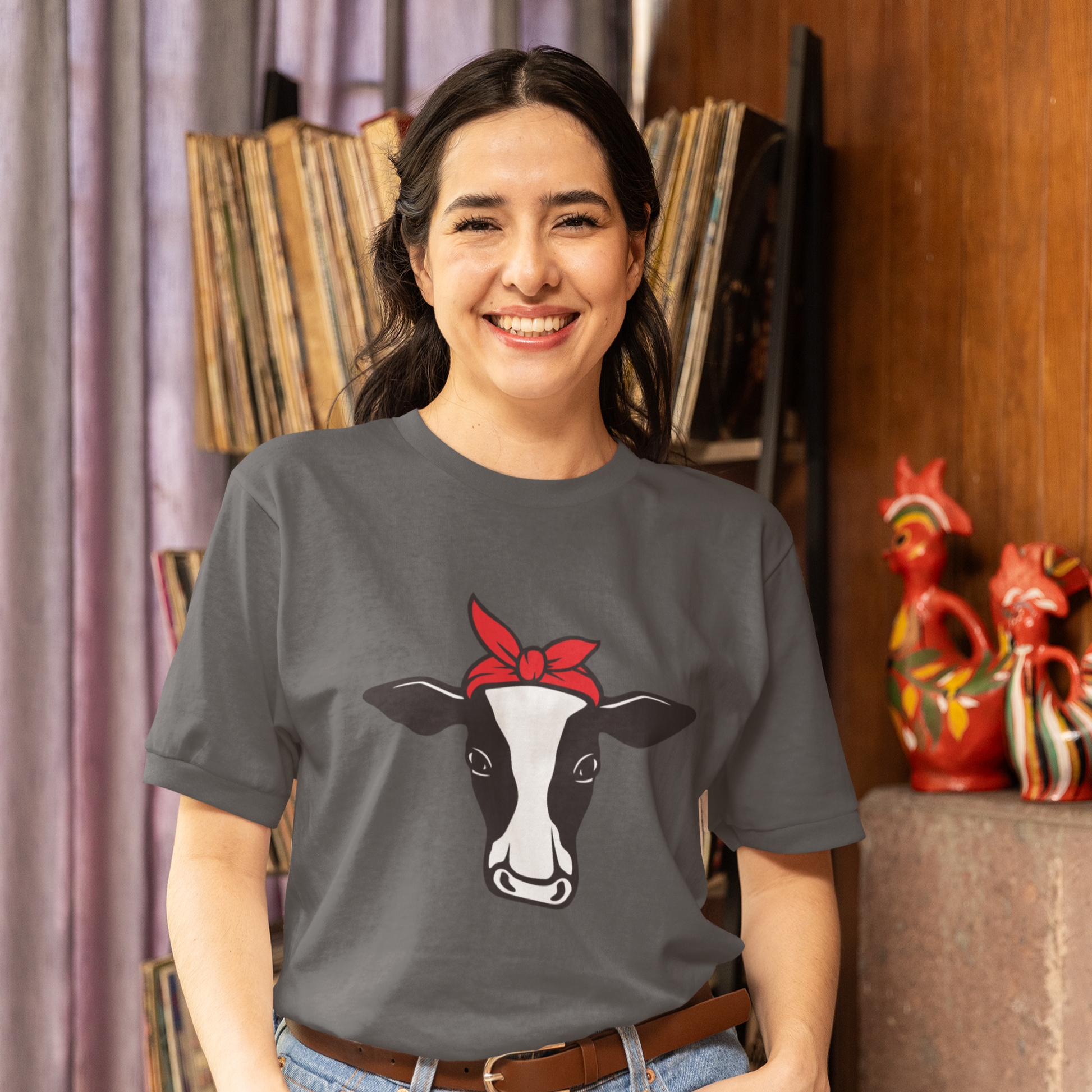 Person wearing a grey t-shirt with a cow graphic and red bandana, standing in a room with books and decorative items.