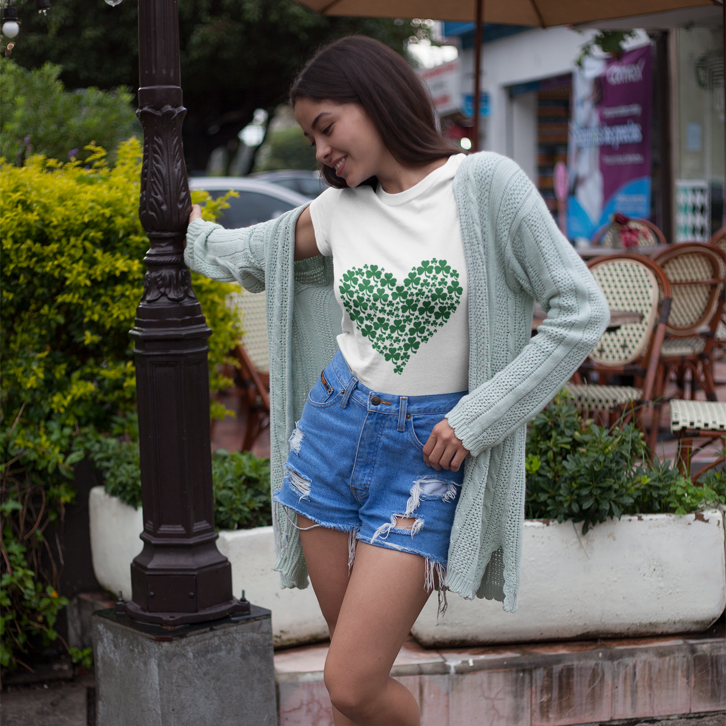 Woman wearing a white t-shirt with a green heart design, light blue cardigan, and denim shorts outdoors.