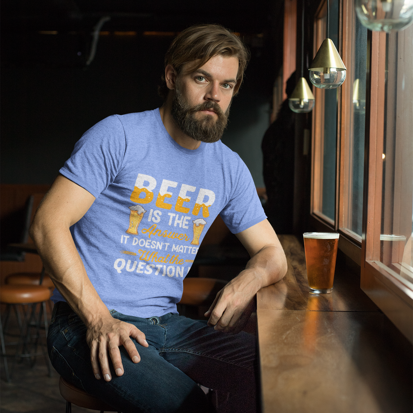 Man sitting at a bar wearing a blue t-shirt with text, holding a beer glass.