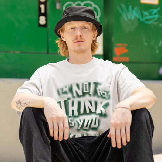 Person sitting on a skateboard wearing a white t-shirt with text and black pants, with a green wall and graffiti in the background.