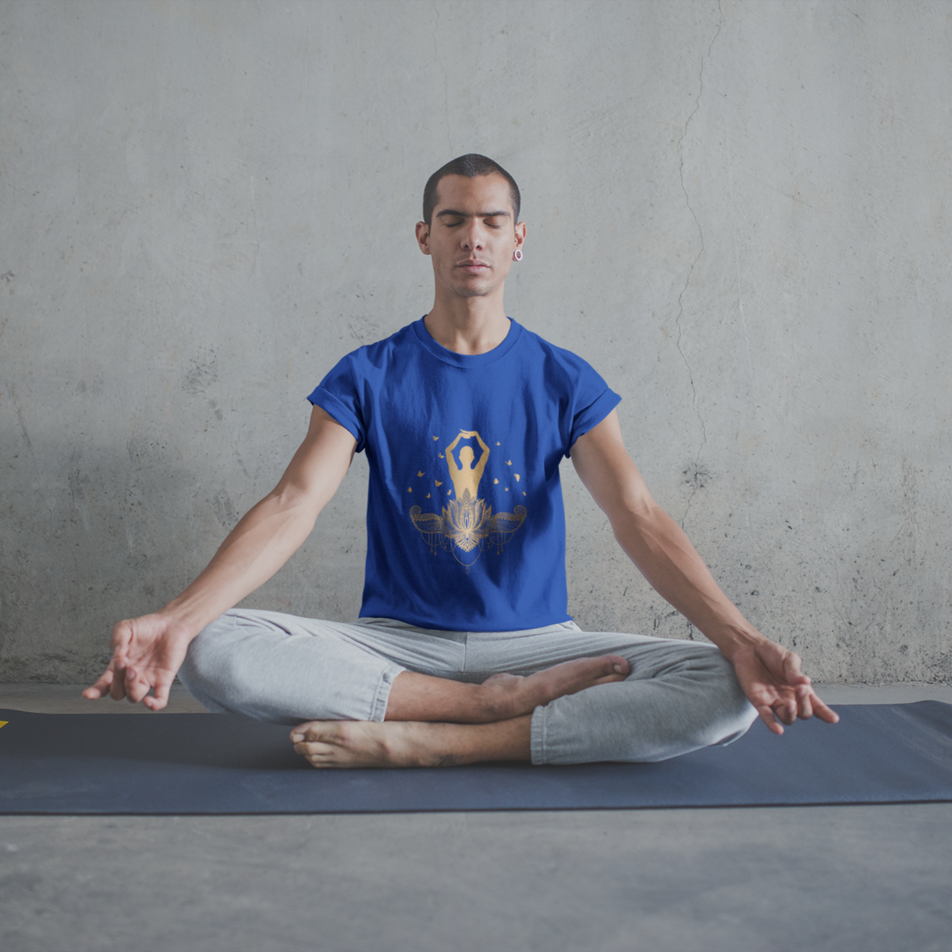 Man in a blue t-shirt and gray pants sitting in a yoga pose on a mat against a plain wall.