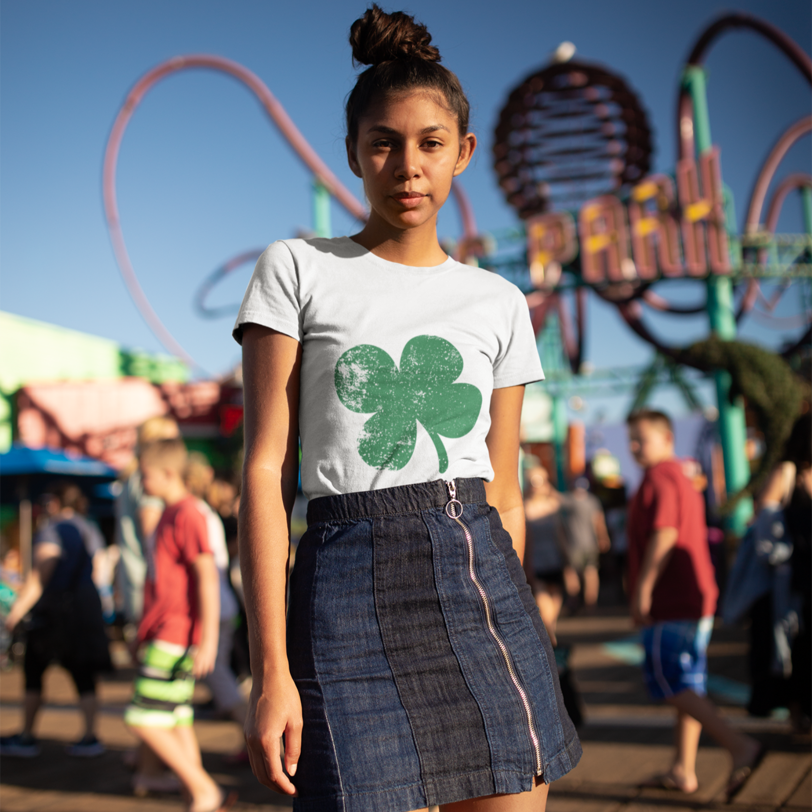 Woman wearing a white t-shirt with a green shamrock design and a denim skirt in an amusement park setting.