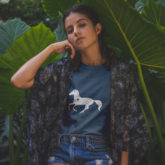 Woman sitting among large green leaves wearing a blue shirt with a horse graphic.
