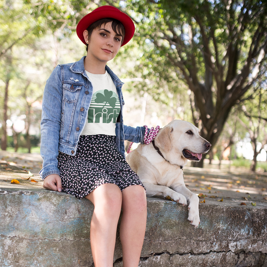 Person sitting on a stone wall with a dog, wearing a red hat, denim jacket, and white shirt with green design.