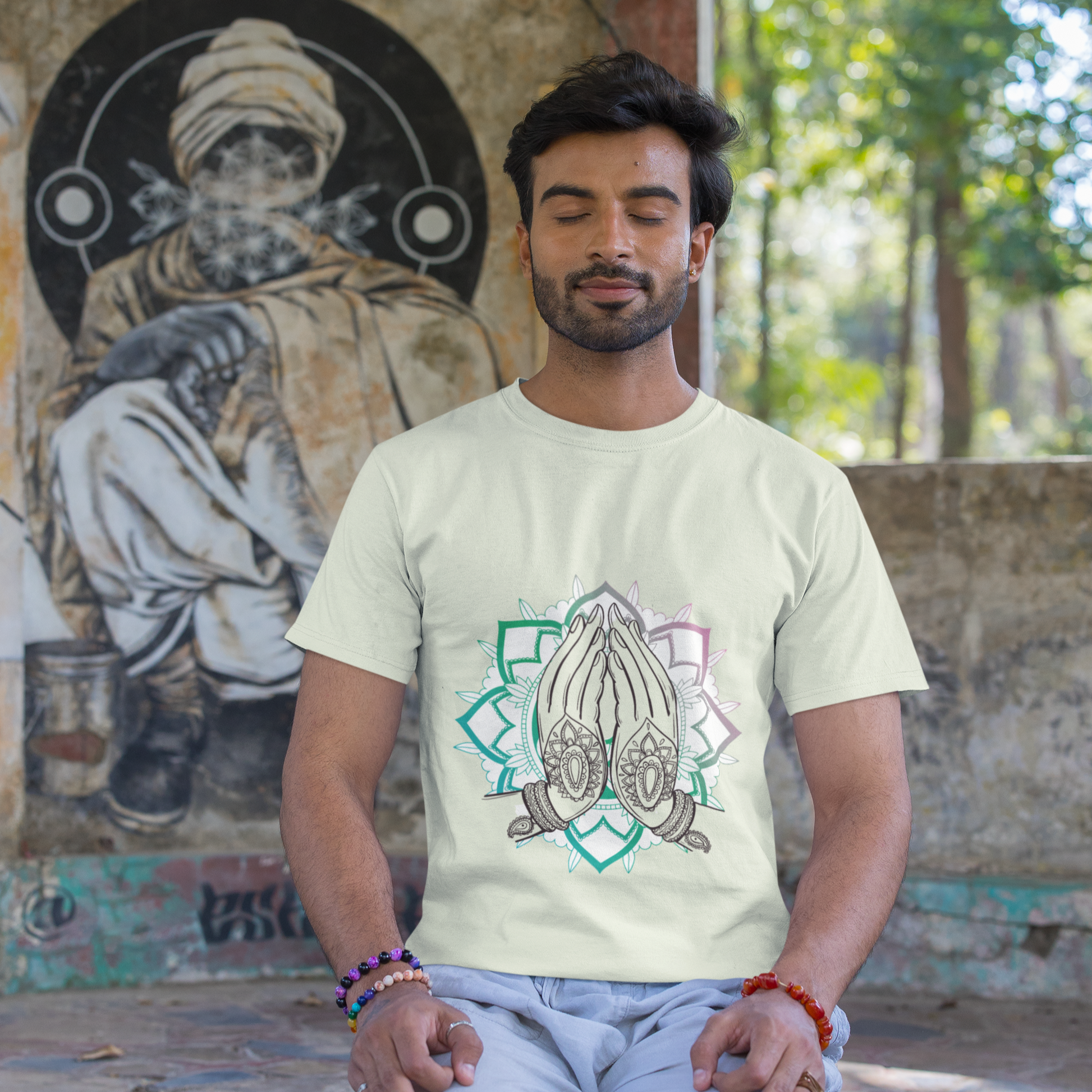 Man sitting on a stone floor wearing a light-colored t-shirt with a design, with a mural in the background.