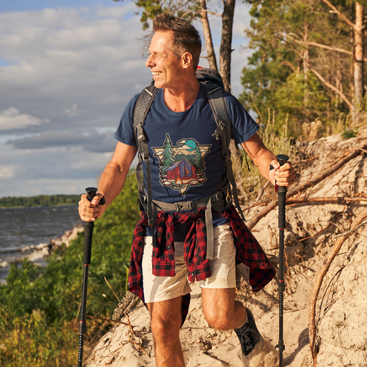 Man hiking on a trail with a backpack and walking sticks, surrounded by trees and water.