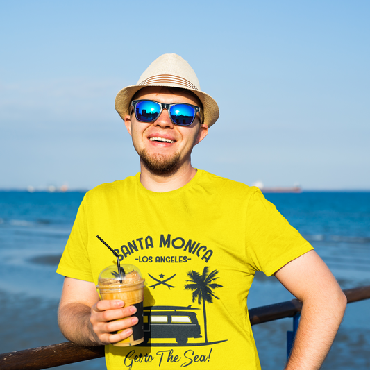 Man wearing a yellow t-shirt with a beach-themed design, sunglasses, and a hat, holding a drink by the ocean.