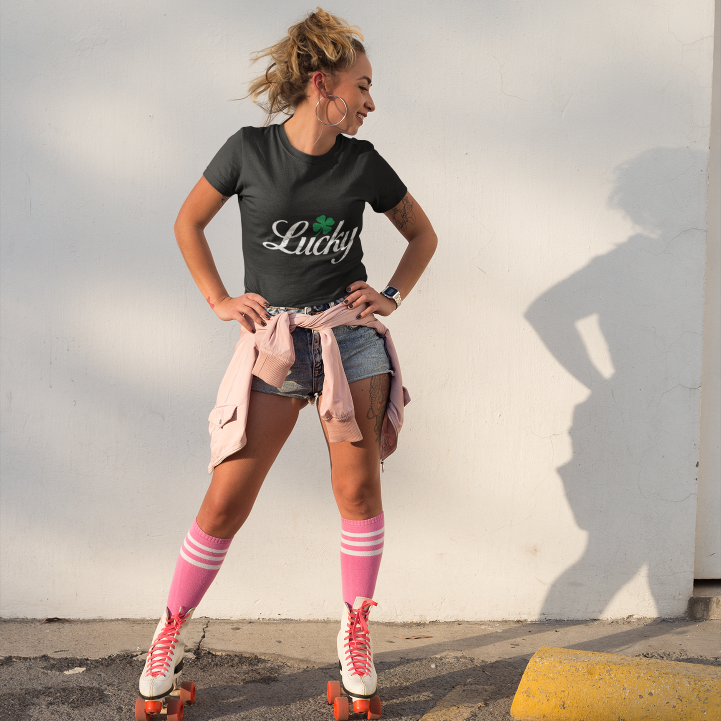 Woman in black t-shirt with 'Lucky' print, pink knee-high socks, and roller skates standing against a light-colored wall.