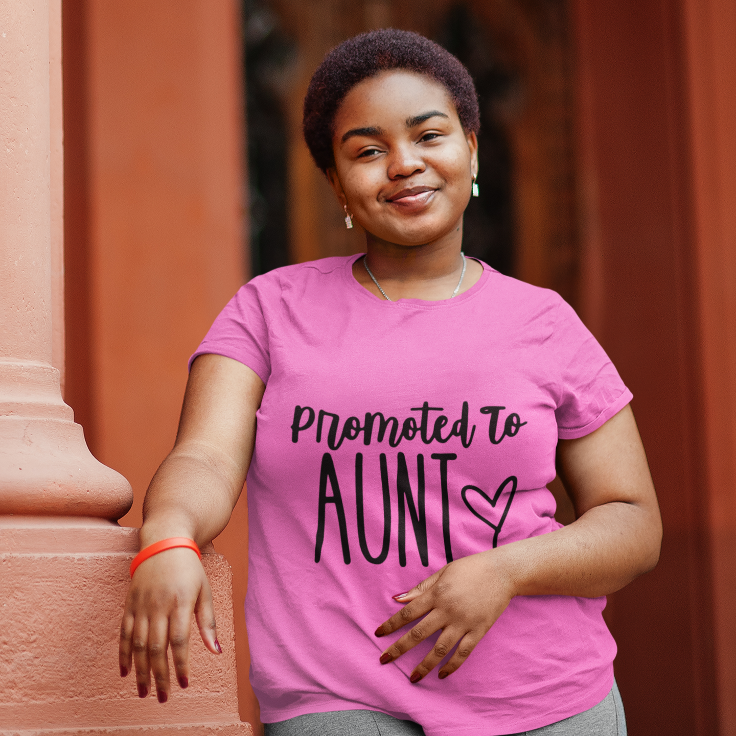 Woman wearing a pink t-shirt with 'Promoted to Aunt' text, standing against a warm-toned wall.