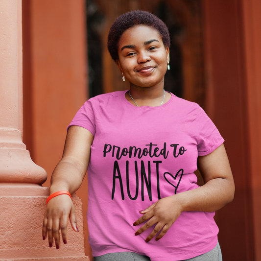 Woman wearing a pink t-shirt with 'Promoted to Aunt' text, standing against a warm-toned wall.