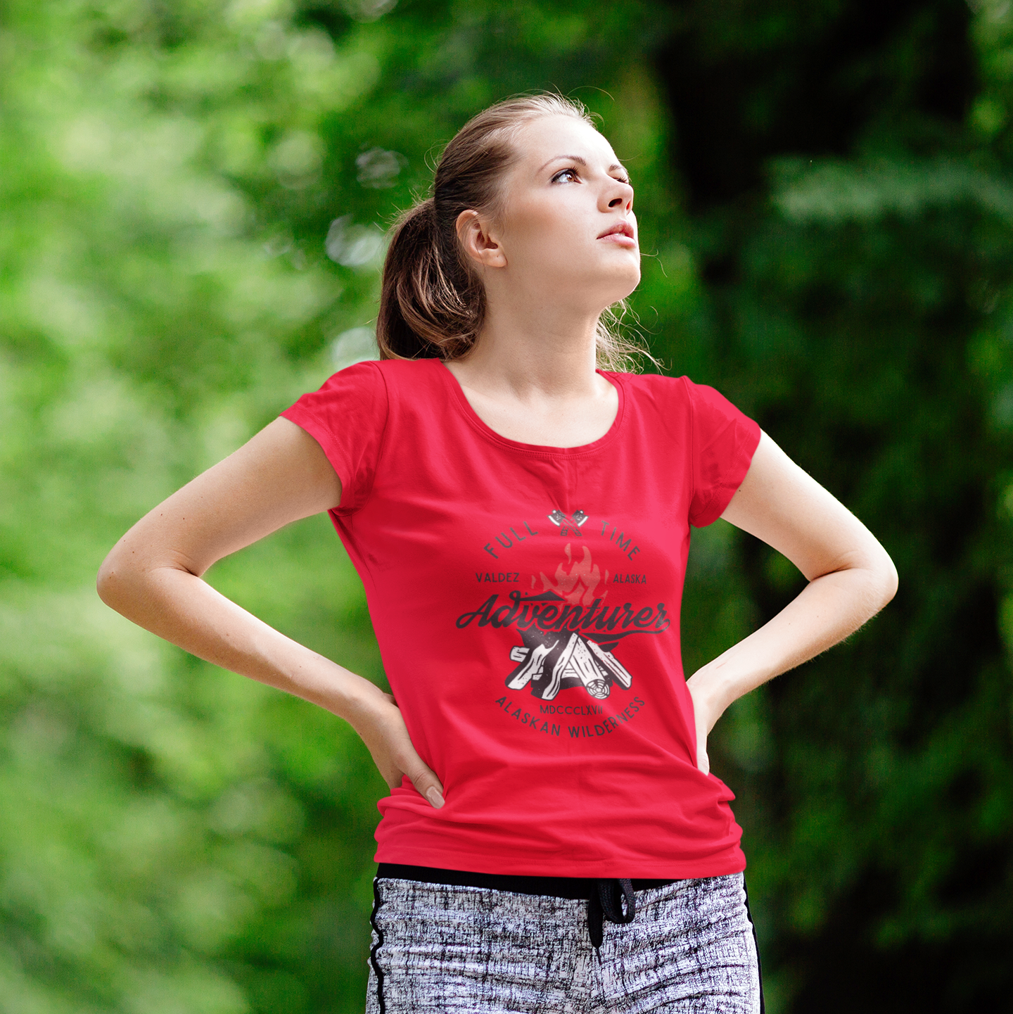 Woman wearing a red t-shirt with text and graphics, standing outdoors with greenery in the background
