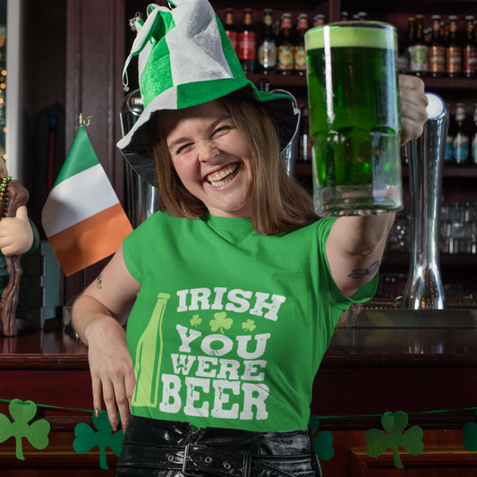 Person wearing an Irish-themed shirt and hat, holding a glass of green beer in a bar setting.
