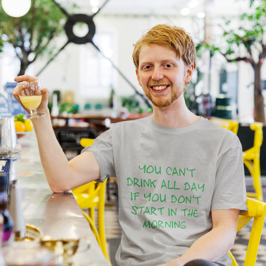Man sitting at a table in a cafe, holding a drink and wearing a t-shirt with a humorous message.