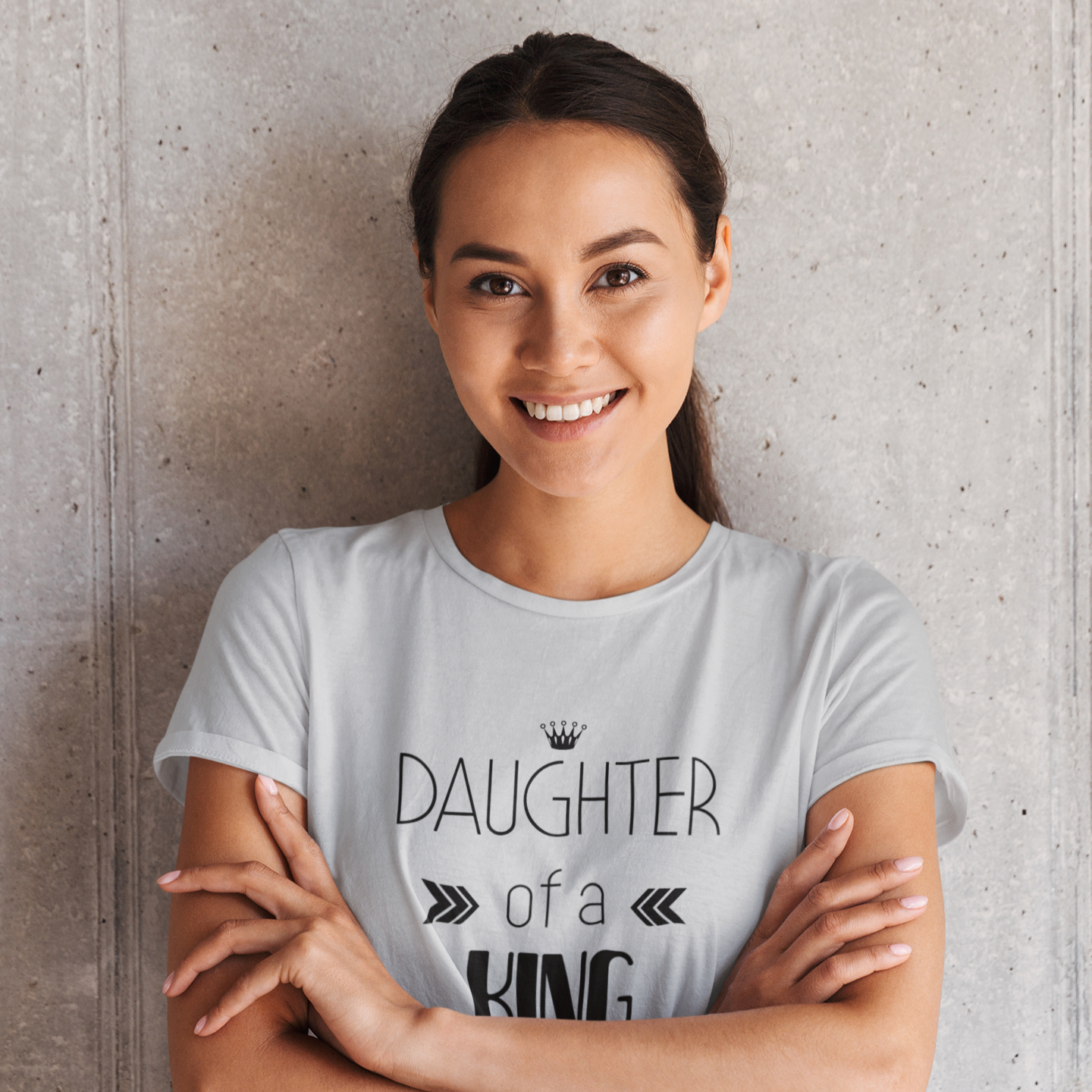 Woman wearing a t-shirt with 'Daughter of a King' text against a concrete wall.