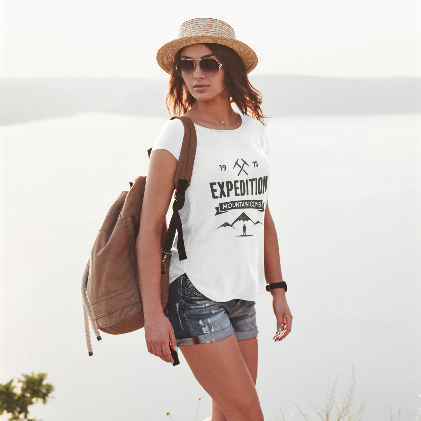 Woman in outdoor setting wearing a white t-shirt with 'Expedition' text, shorts, sandals, and a straw hat.