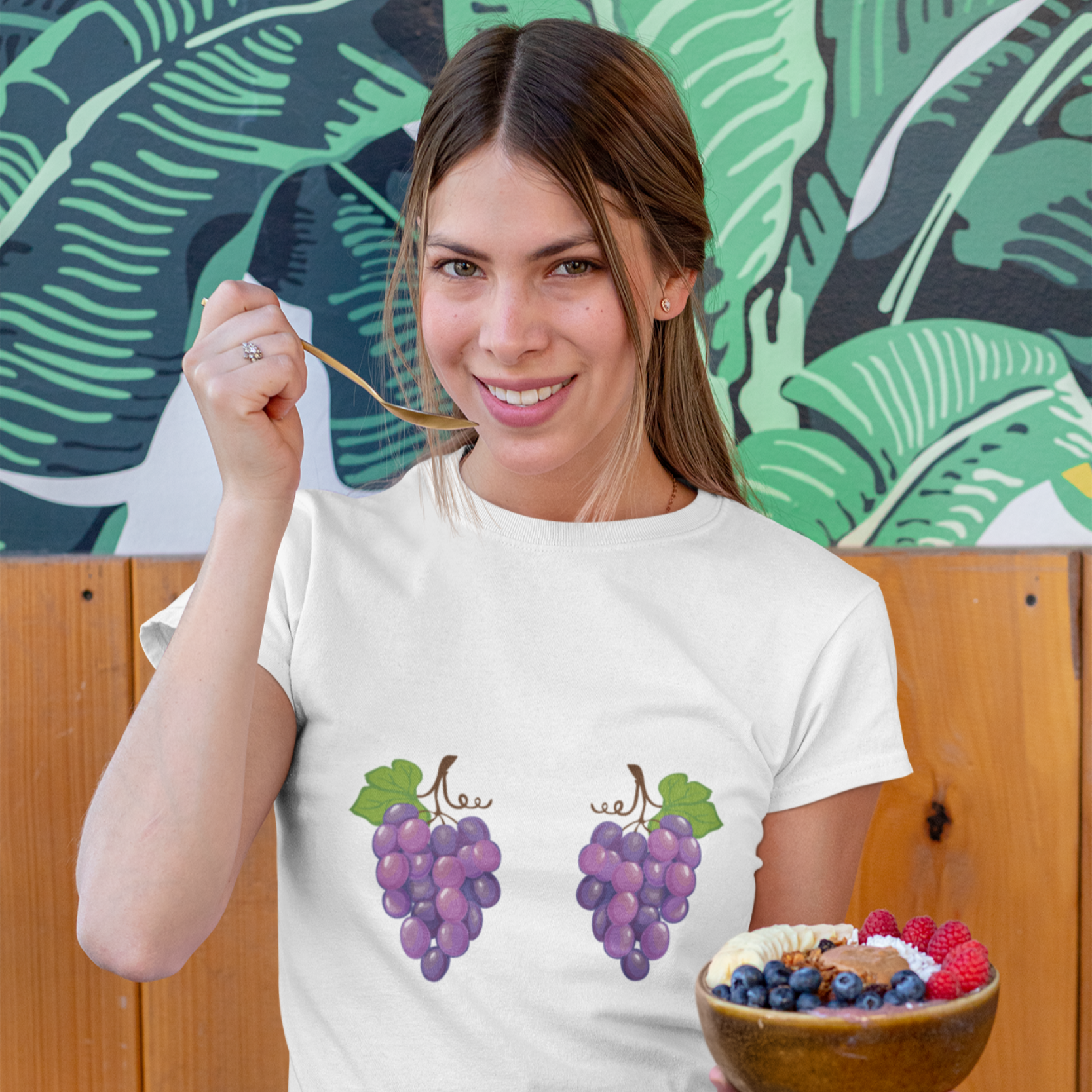Woman wearing a white t-shirt with grape design, holding a bowl of fruit, standing in front of a colorful leafy background.