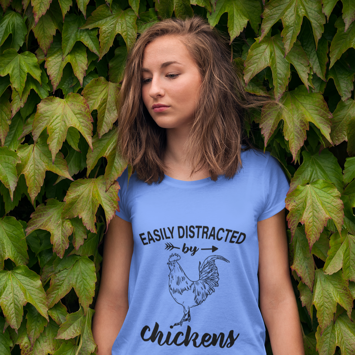 Woman wearing a blue t-shirt with a chicken graphic and text, standing against a leafy background.