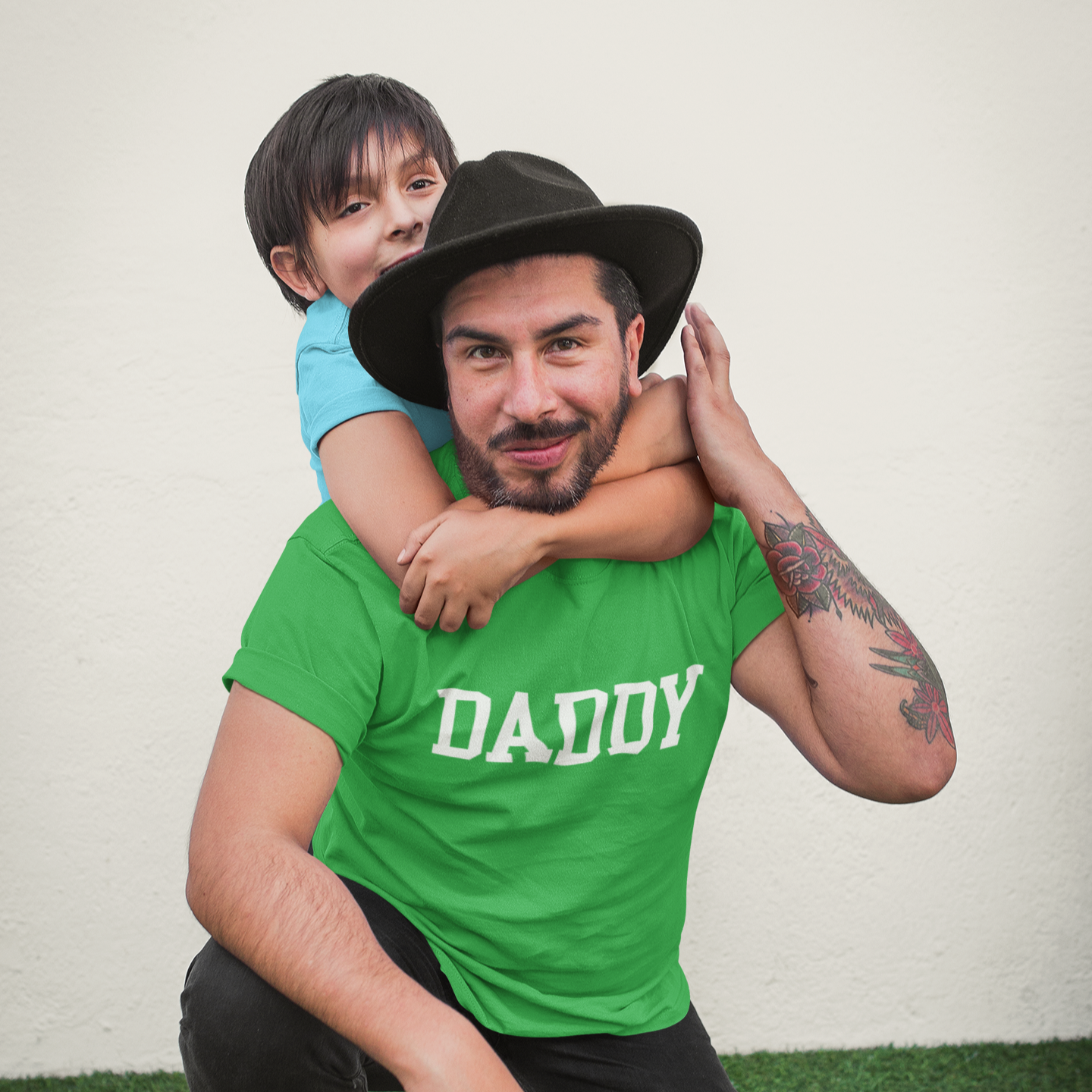 Man wearing a green 'DADDY' shirt holding a child on a grassy area.