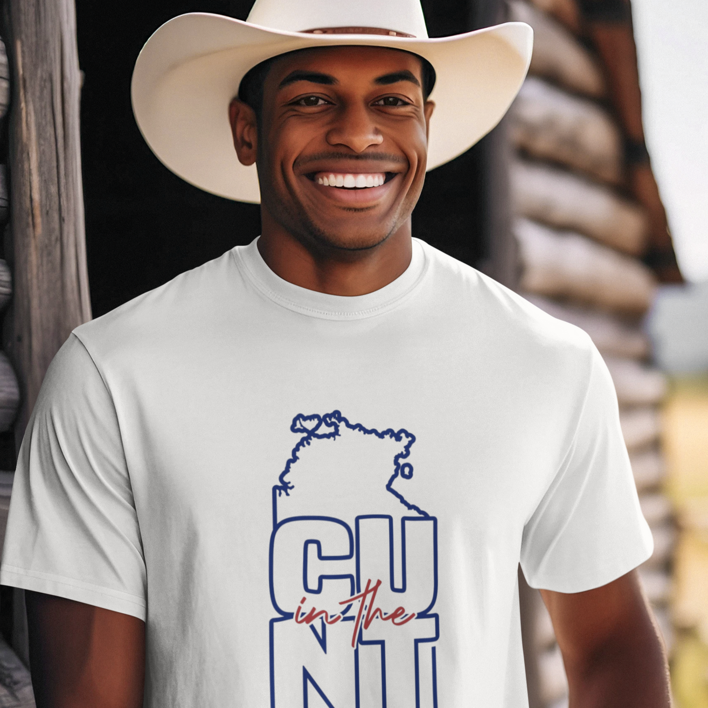 Man wearing a white t-shirt with a graphic design and a cowboy hat, standing in front of a wooden structure.