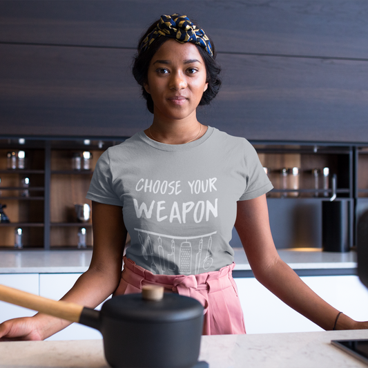 Woman in a kitchen wearing a t-shirt with 'Choose Your Weapon' text