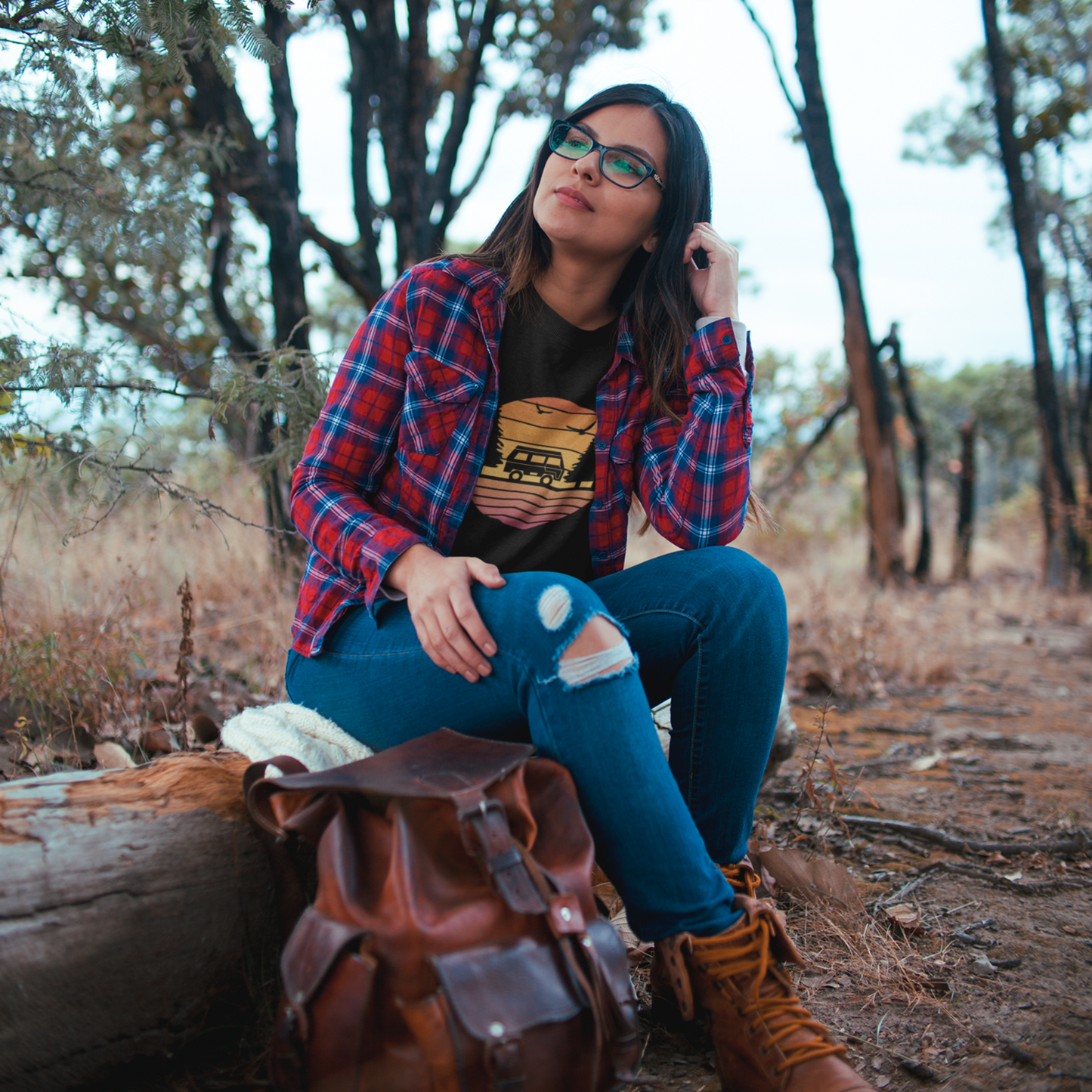 Person sitting outdoors with a backpack, wearing a plaid shirt and jeans.